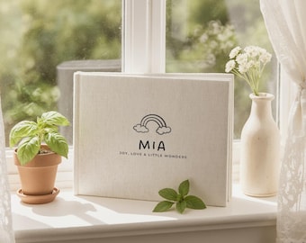 May include: A linen-covered book with the name "MIA" and a rainbow illustration. The text "JOY, LOVE & LITTLE WONDERS" is below the name. A small potted plant and a vase with white flowers sit nearby on a white windowsill.