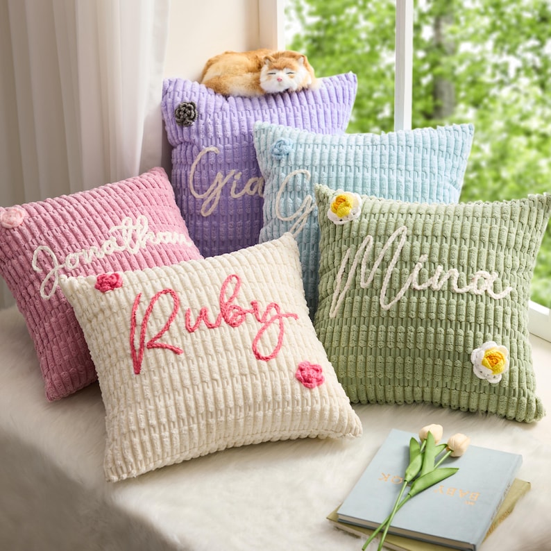 May include: A collection of five square, textured corduroy throw pillows in pastel shades of pink, purple, light blue, cream, and sage green. Each pillow features a personalized name embroidered in white or pink yarn and small floral accents.