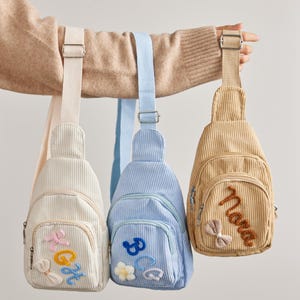 May include: Three corduroy sling bags in cream, light blue, and tan. Each bag features a front zippered pocket with embroidered letters and small decorative bows or flowers. The bags have adjustable woven straps and silver-toned metal hardware.