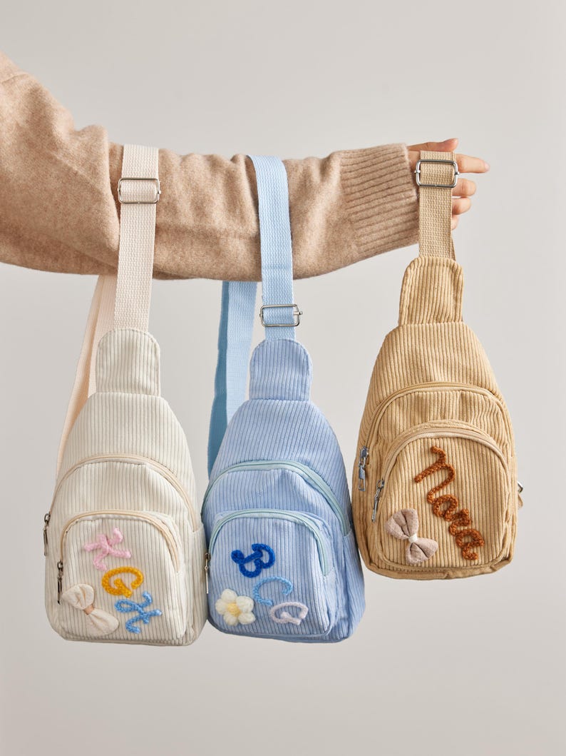 May include: Three corduroy sling bags in cream, light blue, and tan. Each bag features a front zippered pocket with embroidered letters and small decorative bows or flowers. The bags have adjustable woven straps and silver-toned metal hardware.