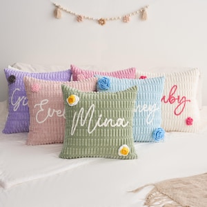 May include: A collection of five square corduroy throw pillows in lavender, dusty pink, sage green, light blue, and cream. Each pillow features a cursive name embroidered in white or pink thread, accented with small, colorful crocheted flower appliqu&eacute;s.