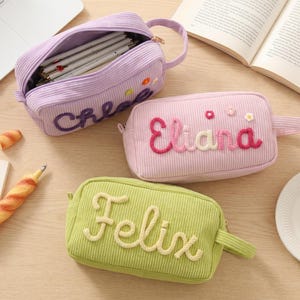 Puede incluir: Tres estuches rectangulares de pana en color lavanda, rosa claro y verde claro. Cada estuche cuenta con un asa lateral, cierre de cremallera y un nombre bordado en letras con relieve y textura: Chloe, Eliana y Felix.