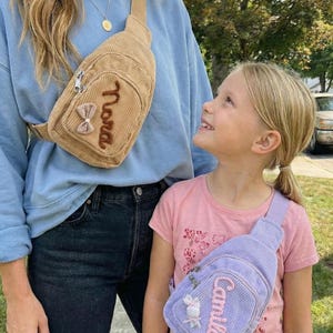 May include: A tan corduroy crossbody bag with a brown embroidered name and a small bow, and a light purple corduroy crossbody bag with a pink embroidered name and a small bow. Both bags feature a zippered front pocket and an adjustable strap.