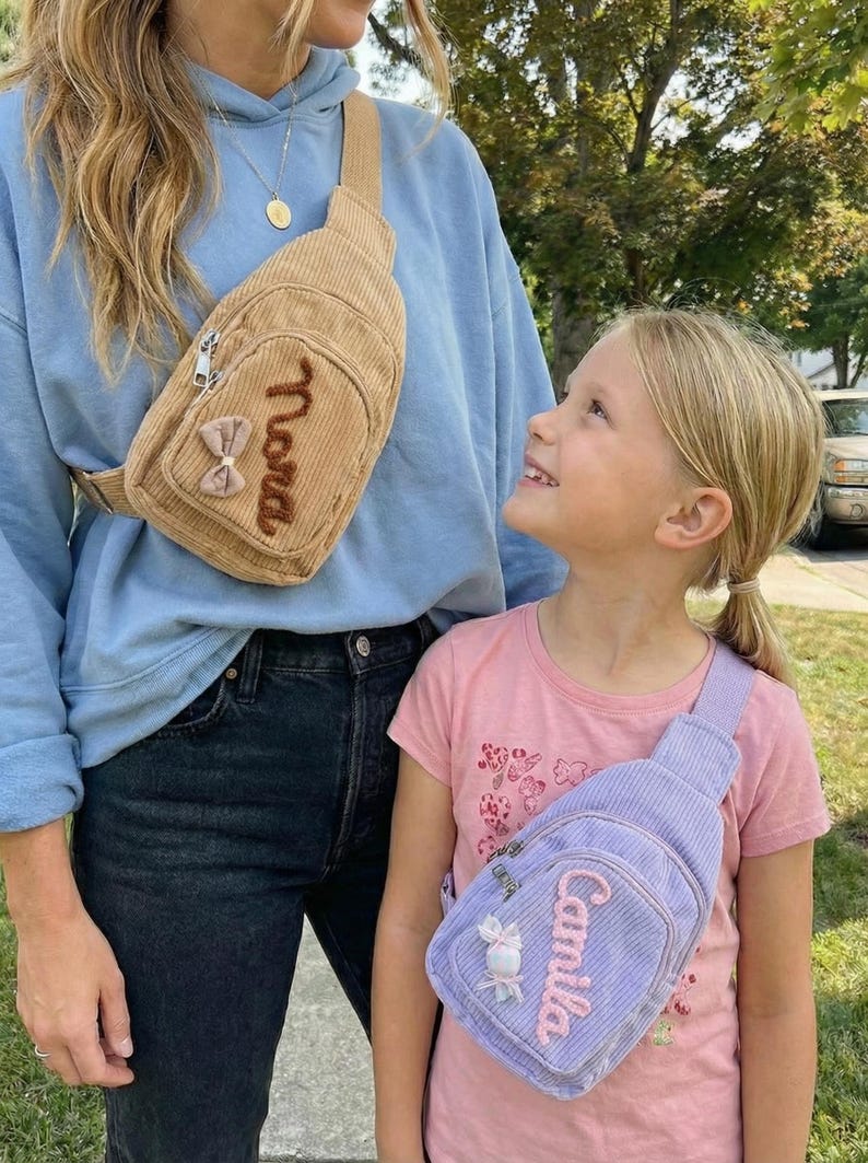May include: A tan corduroy crossbody bag with a brown embroidered name and a small bow, and a light purple corduroy crossbody bag with a pink embroidered name and a small bow. Both bags feature a zippered front pocket and an adjustable strap.