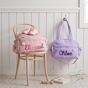 May include: A pink duffel bag with the name Eliana embroidered in pink and white, featuring a small white flower and rabbit charm, sits on a wooden chair with pink trainers. A purple duffel bag with the name Chloe embroidered in purple.