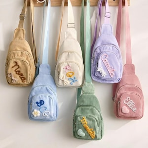 May include: A collection of six corduroy sling bags in tan, light blue, cream, green, lavender, and pink, each featuring a front zippered pocket with embroidered names and small decorative appliques like bows, flowers, and hearts.