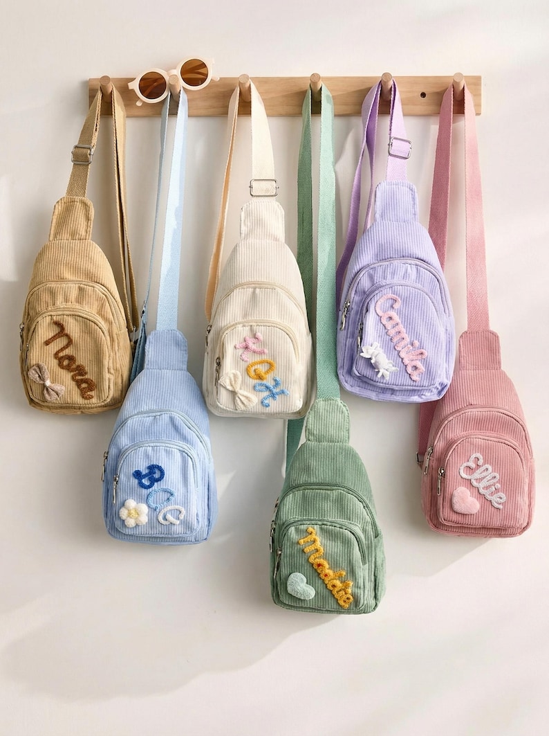 May include: A collection of six corduroy sling bags in tan, light blue, cream, green, lavender, and pink, each featuring a front zippered pocket with embroidered names and small decorative appliques like bows, flowers, and hearts.