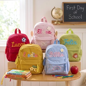 May include: Five corduroy backpacks in red, pink, yellow, light blue, and lime green. Each bag features a personalized name in embroidered script, a top handle, a front zippered pocket, and unique patches like a soccer ball, bow, flower, or rainbow.