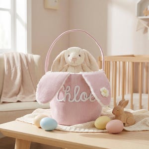 以下が含まれることがあります： ピンク色のコーデュロイのイースターバスケット。ウサギの耳のアクセント、前面に刺繍された「Chloe」の文字、そして中にぬいぐるみウサギが入っています。装飾用の卵と木製のウサギが、クリーム色の毛布の上に置かれています。