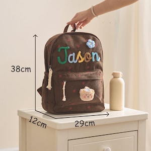 Op de afbeelding: Een bruine corduroy rugzak met de naam "Jason" geborduurd in groen, geel en blauw. De rugzak is 38 cm hoog, 29 cm breed en 12 cm diep. Hij heeft een kleine berenpatch en een blauwe bloem.