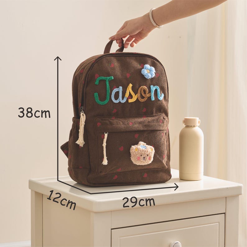 Op de afbeelding: Een bruine corduroy rugzak met de naam "Jason" geborduurd in groen, geel en blauw. De rugzak is 38 cm hoog, 29 cm breed en 12 cm diep. Hij heeft een kleine berenpatch en een blauwe bloem.