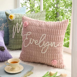 May include: A square throw pillow in dusty pink with a textured corduroy-like surface. The name Evelyn is embroidered in white cursive script across the center. Two small, light pink crocheted flowers are attached to the top and bottom corners.