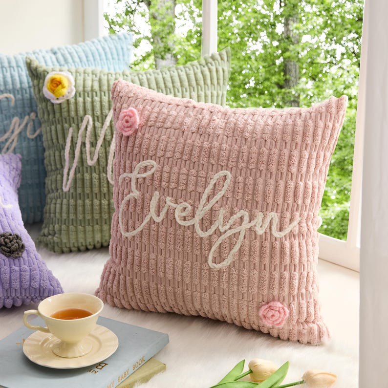 May include: A square throw pillow in dusty pink with a textured corduroy-like surface. The name Evelyn is embroidered in white cursive script across the center. Two small, light pink crocheted flowers are attached to the top and bottom corners.