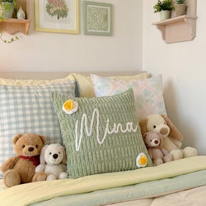 May include: A sage green square throw pillow with a textured corduroy-like surface, featuring the name Mina embroidered in white cursive script and two small white and yellow flower appliqu&eacute;s on opposite corners, resting on a bed.
