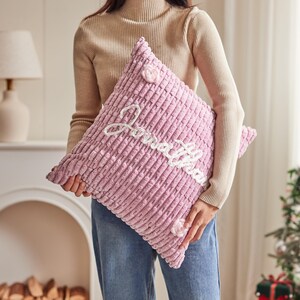 May include: A square, light pink, textured corduroy throw pillow featuring the name Jonathan embroidered in white cursive script across the center, accented with two small, light pink crocheted flower embellishments on the top and bottom.