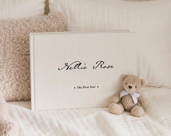 May include: A cream-colored baby photo album with the name "Nellie Rose" in script and the text "The First Year." A small teddy bear with a blue bow sits next to the album on a white bed.