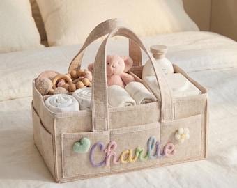 May include: A beige felt diaper caddy with two handles, featuring the name Charlie embroidered in pastel yarn on the front. The organizer includes a small green heart and white flower applique, filled with baby items like a plush toy and wipes.