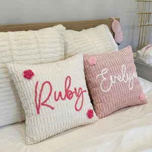 May include: Two square throw pillows with a textured, ribbed fabric. One white pillow features the name Ruby in pink script with two small pink crocheted flowers. One dusty pink pillow features the name Evelyn in white script with two small pink flowers.