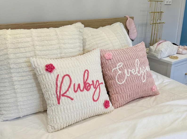 May include: Two square throw pillows with a textured, ribbed fabric. One white pillow features the name Ruby in pink script with two small pink crocheted flowers. One dusty pink pillow features the name Evelyn in white script with two small pink flowers.