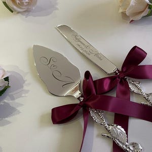 Può includere: Set di coltello e paletta per torta argentati con nastro bordeaux e decorazioni a rosa. La paletta è incisa con "S & A" e il coltello con "Sufiyan & Alisha 06.01.2026". Perfetto per matrimoni e occasioni speciali.