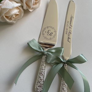 Può includere: Set di coltello e paletta per torta argentati con manici floreali incisi e fiocchi di nastro verde salvia. Il server è inciso con "Samuel & Rachel 11.07.2026". Il coltello ha le iniziali "SR". Set per servire la torta nuziale.