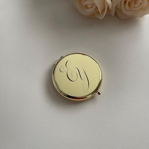 Puede incluir: Un espejo de bolsillo redondo de metal dorado con una superficie pulida y reflectante. La parte superior del estuche está grabada con las letras cursivas entrelazadas "Ey" en una caligrafía fina y oscura, con una pequeña bisagra.