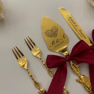May include: A gold-coloured wedding cake serving set, including a cake server, knife, and two forks. The server and knife are engraved with names and a quote. The forks are engraved with "Mr" and "Mrs". Tied with a burgundy ribbon.