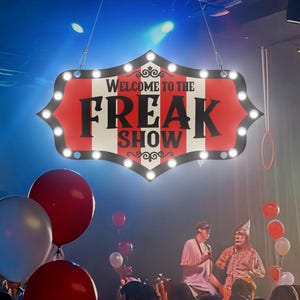 以下が含まれることがあります： 「Welcome to the Freak Show」と書かれた看板が人々の頭上に吊り下げられています。看板は赤と白で、装飾的な縁とライトが付いています。赤い風船と白い風船が前景に浮かんでおり、2人のパフォーマーがステージにいます。