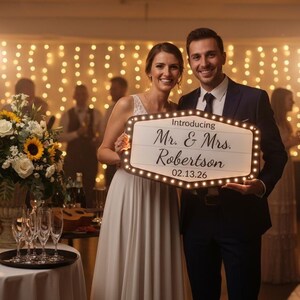 May include: A couple smiles, holding a sign that reads "Introducing Mr. & Mrs. Robertson 02.13.26." The sign is a light-up marquee with a wooden frame. The woman wears a white dress, and the man wears a navy suit.