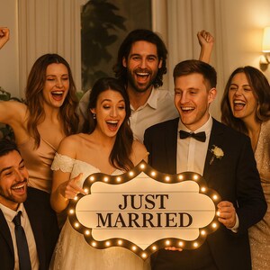 May include: A group of people celebrating, holding a lighted sign that reads "JUST MARRIED". The sign has a wooden background with a decorative border of small lights. The people are smiling and laughing, dressed in formal attire.