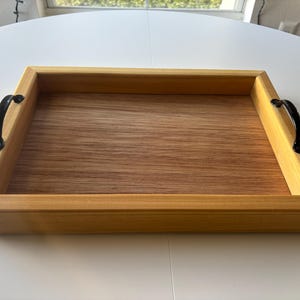 May include: A rectangular wooden serving tray with a light brown interior and a medium brown frame. The tray has black metal handles on each end. The wood grain is visible on the tray's surface.