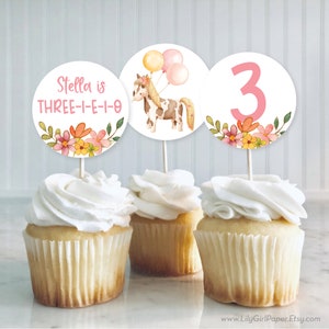 May include: Three cupcake toppers with a pink and white floral design. One topper features a brown and white pony with pink balloons, another says "Stella is THREE-E-I-O", and the last one has a pink number 3.