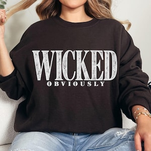May include: A dark brown crewneck sweatshirt featuring the word WICKED in large, distressed white capital letters, with the word OBVIOUSLY printed in a smaller, clean white font directly underneath the main text on the front of the garment.