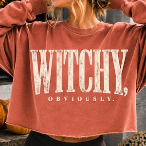 May include: A rust-colored cropped sweatshirt with long sleeves and a raw-edge hem. The front features the distressed white text "WITCHY," in a large serif font, with the word "OBVIOUSLY." printed in a smaller, matching font directly below it.
