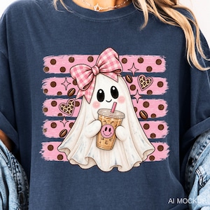 May include: A dark blue t-shirt featuring a graphic of a white ghost wearing a pink gingham bow, holding an iced coffee cup with a pink smiley face. The background includes pink horizontal brush strokes, brown polka dots, hearts, and coffee beans.