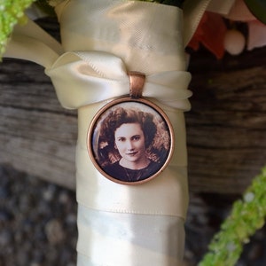 May include: A white satin ribbon bouquet with a copper pendant featuring a black and white photo of a woman.