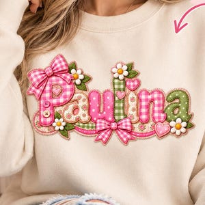 Könnte beinhalten: Ein cremefarbenes Sweatshirt mit Rundhalsausschnitt und dem Namen Paulina in einer gemusterten, gesteppten Schriftart. Das Design umfasst rosa und grünes Vichy-Karo, florale Muster, aufgenähte Schleifen, kleine Herzen und weiße Gänseblümchen.