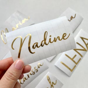 Könnte beinhalten: Eine Sammlung von personalisierten Vinyl-Aufklebern mit Namen in metallisch goldener Schreibschrift. Jeder Aufkleber befindet sich auf einem weißen Transferpapier mit Gittermuster, darunter die Namen Nadine, Lena und weitere.