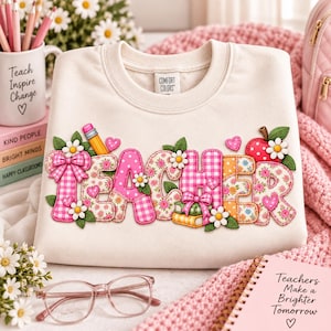 Peut inclure: Un sweat-shirt à col rond couleur crème portant le mot TEACHER en grandes lettres gonflées à motifs roses, décorées de marguerites, de cœurs, d'un crayon et d'une pomme. Le tissu présente une bordure cousue visible autour de chaque lettre.