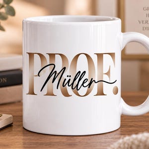 Könnte beinhalten: Eine weiße Keramiktasse mit dem Text PROF. in einer großen, metallisch bronzefarbenen Verlaufs-Schriftart, wobei der Name Müller in einer schwarzen Schreibschrift mittig darüber platziert ist. Die Tasse hat einen Henkel.