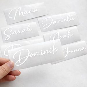 Könnte beinhalten: Weiße Namensaufkleber in Schreibschrift. Die Namen Maria, Daniela, Sarah, Noah, Dominik und Jounna sind zu sehen. Die Aufkleber befinden sich auf einem karierten Hintergrund.