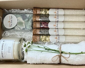 Puede incluir: Un set de regalo de spa en una caja de cartón marrón con papel de seda blanco. El set incluye una toalla blanca, una vela en un frasco de vidrio etiquetado como "Sweet Vanilla", una barra de jabón etiquetada como "The Zen Spa", y seis tubos de vidrio llenos de sales de baño etiquetados como "The Zen ghe Zen Zen Sithe Zen Zen Sp".