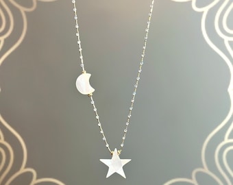 Puede incluir: Un delicado collar con un colgante de luna creciente y una estrella de cinco puntas. Los colgantes son blancos y parecen estar hechos de nácar. La cadena es dorada con pequeñas cuentas transparentes. El collar se muestra sobre un fondo gris.