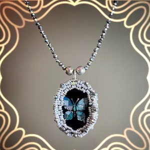 Puede incluir: Un collar de cadena de plata con un colgante ovalado de cuentas que presenta una mariposa azul y verde sobre un fondo negro.
