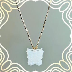 Puede incluir: Un delicado collar con un colgante de mariposa. El colgante es blanco e intrincadamente tallado, suspendido de una cadena de cuentas doradas y plateadas. El collar se muestra sobre un fondo suave y estampado.