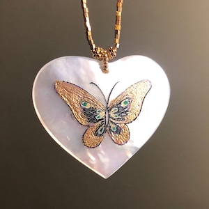 Puede incluir: Un collar de cadena dorada con un colgante en forma de corazón hecho de nácar. El colgante presenta una mariposa pintada con alas doradas y negras y detalles verdes.