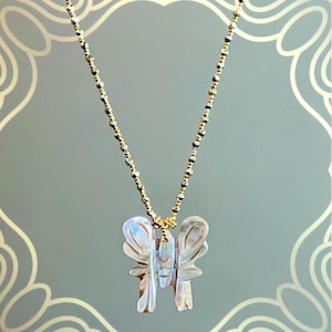 Puede incluir: Un collar de cadena dorada con un colgante de mariposa. El colgante está tallado en un material nacarado, mostrando un diseño de mariposa. El collar es una joya delicada.
