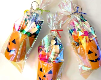 May include: Three Halloween-themed gift bags. Each bag features an orange pumpkin-shaped container filled with treats and wrapped in iridescent cellophane. The bags are tied with ribbons and keychains.