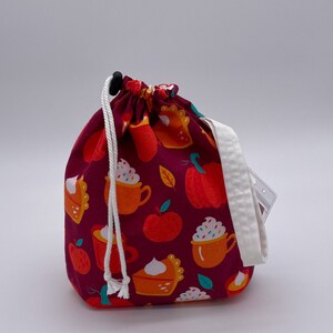 May include: A red drawstring bag with a white cord and a white trim. The bag is decorated with a pattern of orange pumpkins, apple slices, and cups of hot chocolate with whipped cream.