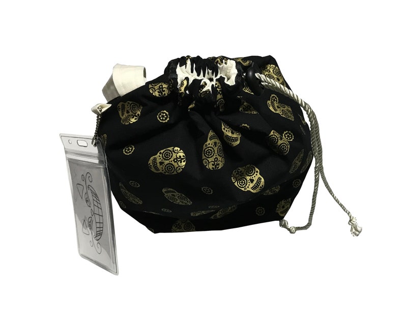 May include: Black drawstring bag with a gold sugar skull pattern. The bag has a white drawstring and a clear plastic ID holder attached.